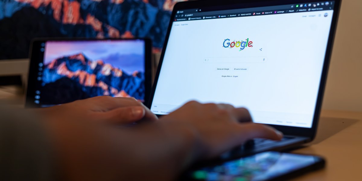 Hands typing on a laptop showing the Google homepage, with a tablet and a smartphone on the desk