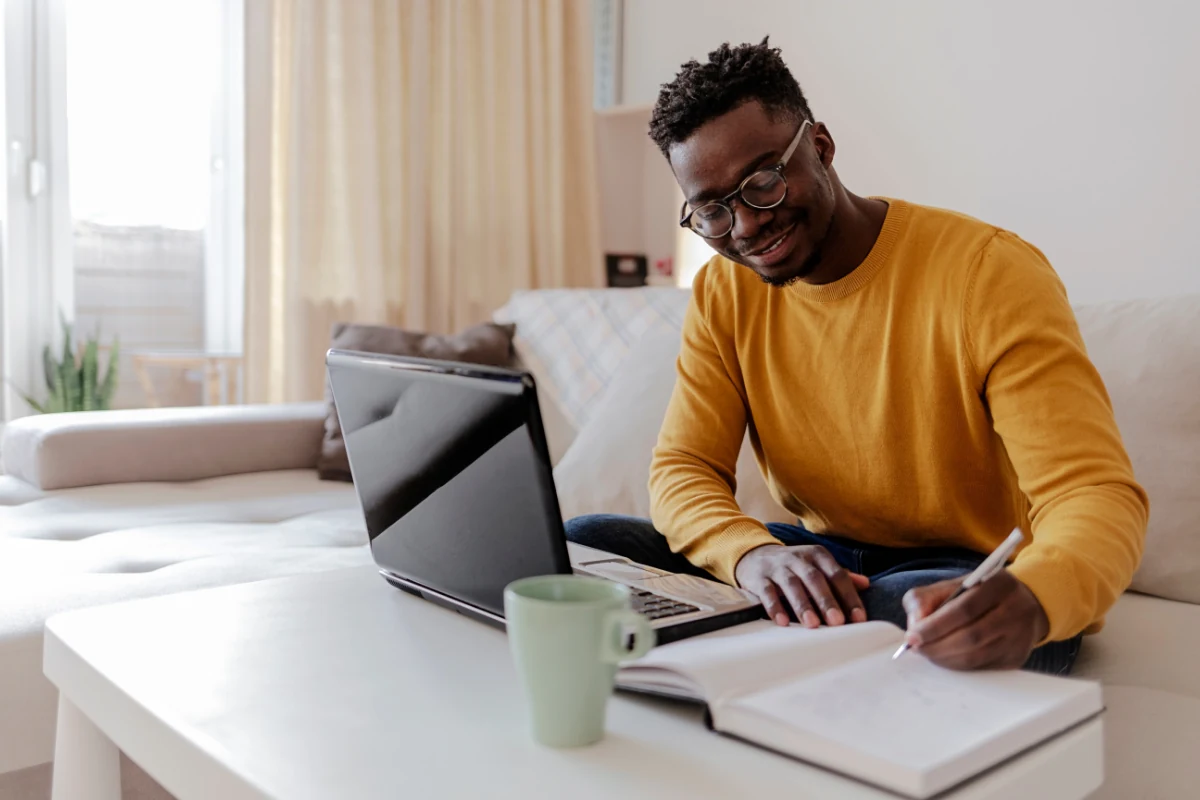 Man on a sofa writing in a notebook next to his laptop and a mug