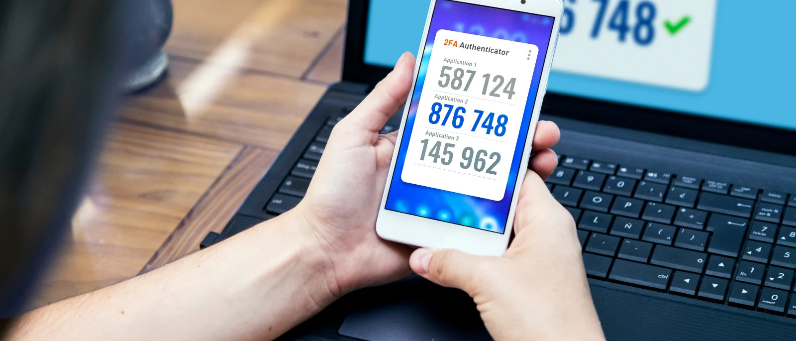 Hands holding a phone showing two-factor authentication codes in front of a laptop