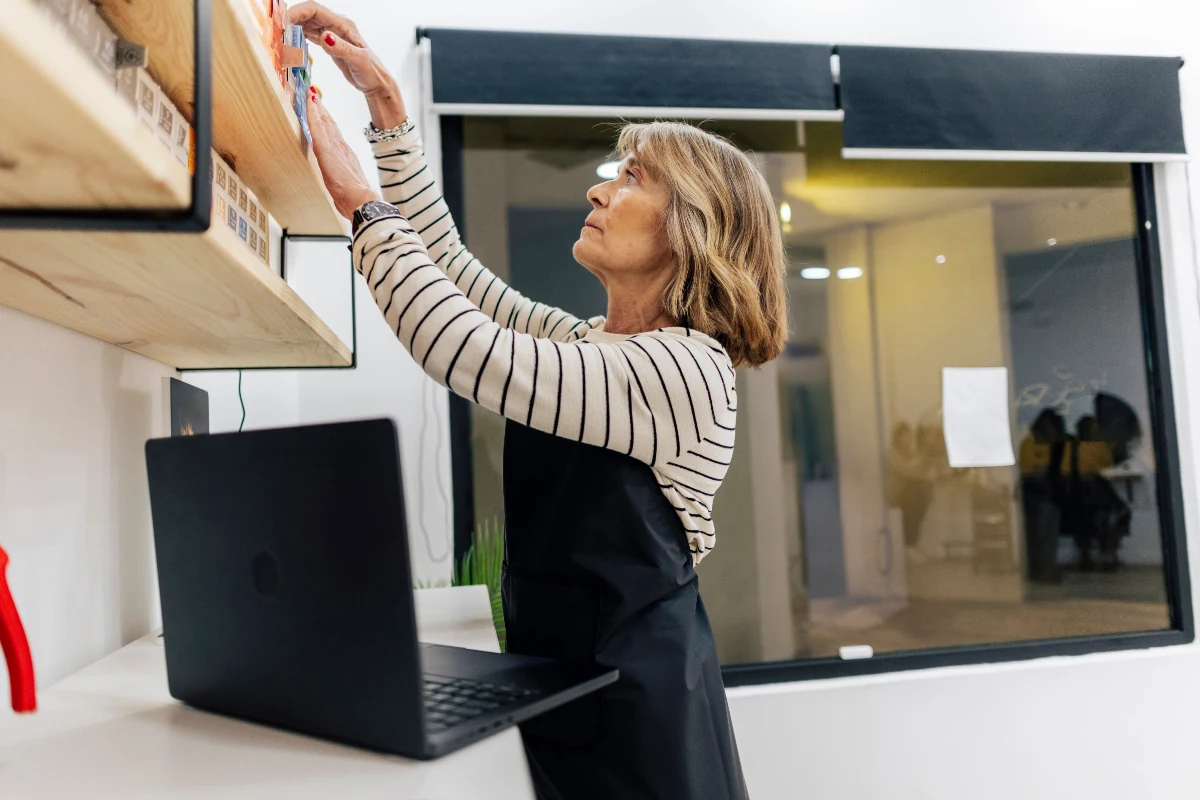 Shop owner stocking a wall shelf in her small store, with a laptop open on the counter