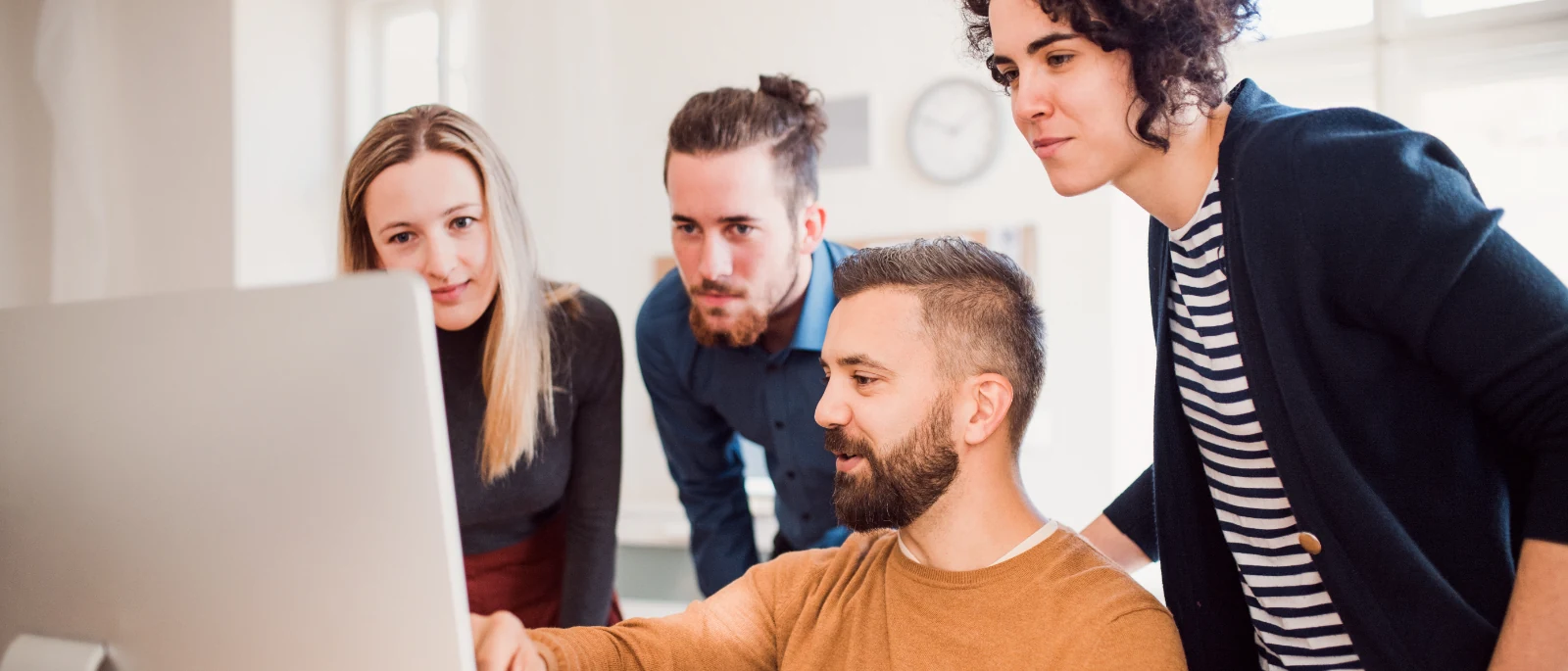 Four colleagues gathered around one desktop monitor while one of them points at the data on screen