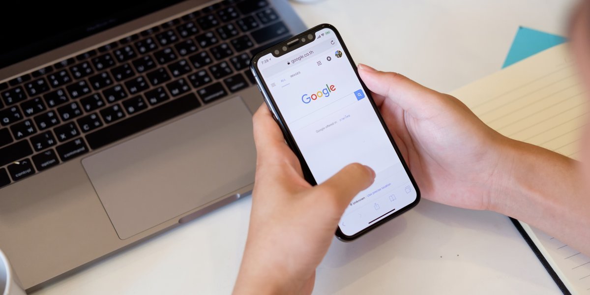 Hands holding a smartphone with the Google search page open, a laptop on the desk behind